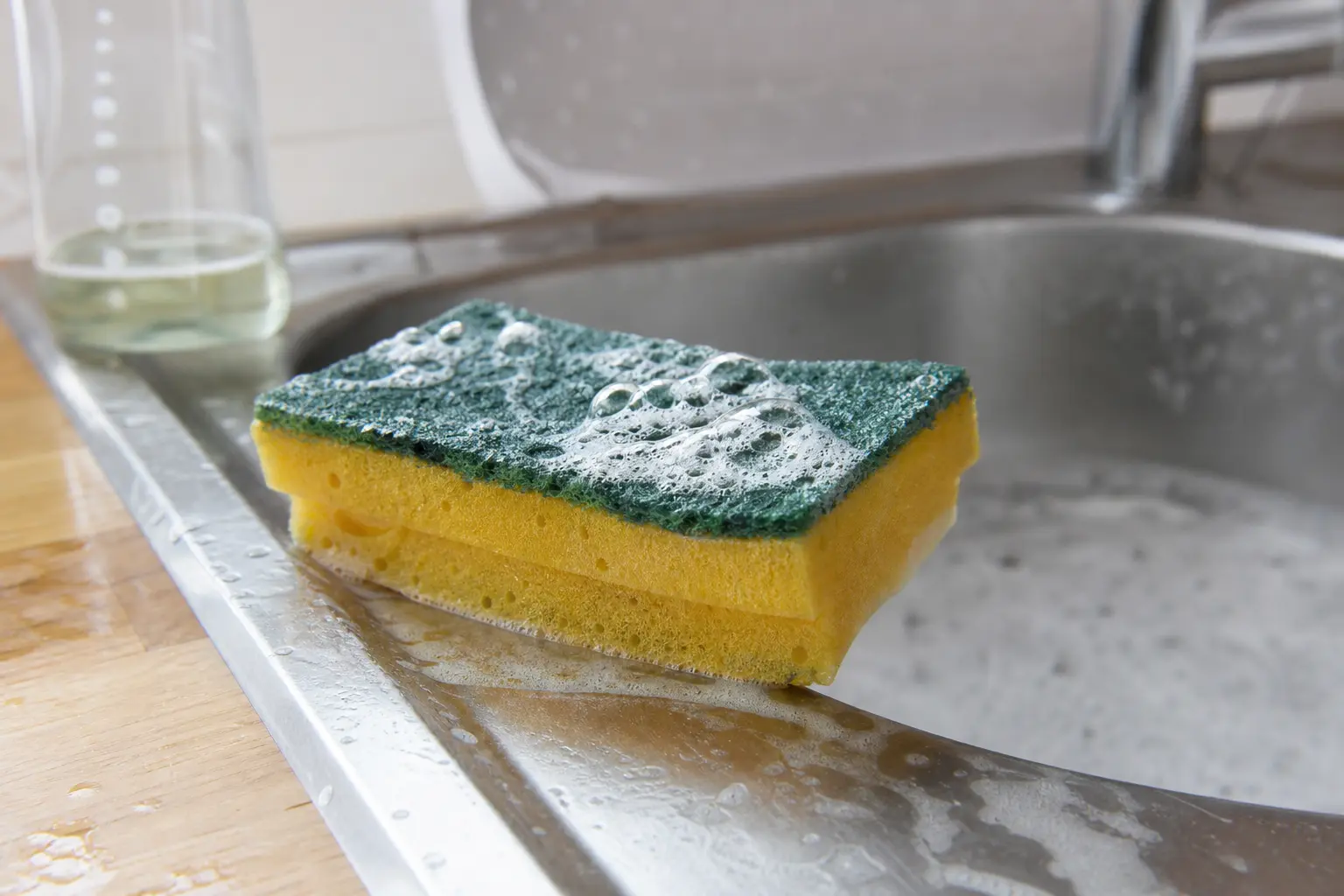 Éponge de cuisine jaune et verte posée sur le bord d'un évier en inox, recouverte de mousse savonneuse, lumière naturelle