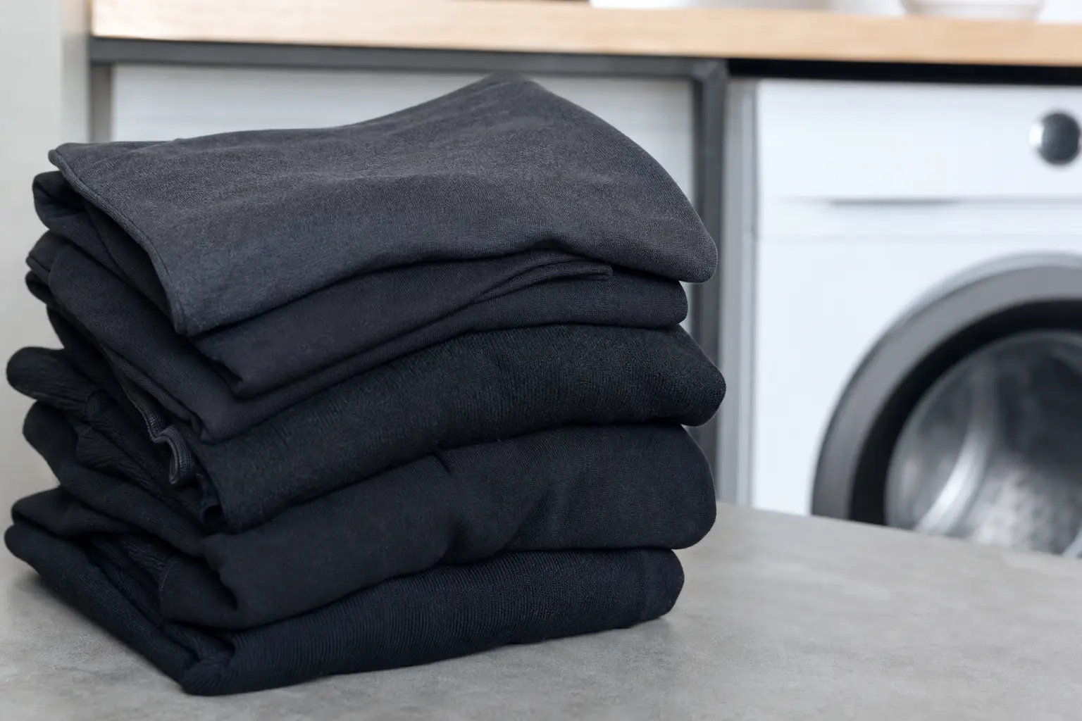 Pile de vêtements noirs pliés près d'un lave-linge dans une buanderie lumineuse, légère nuance grisée visible sur certaines pièces